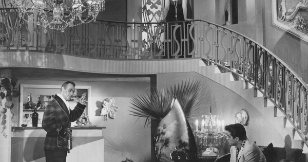 A film still showing two men sitting in a foyer. There is a large curving staircase. One man gesticulates emphatically while speaking to the other.