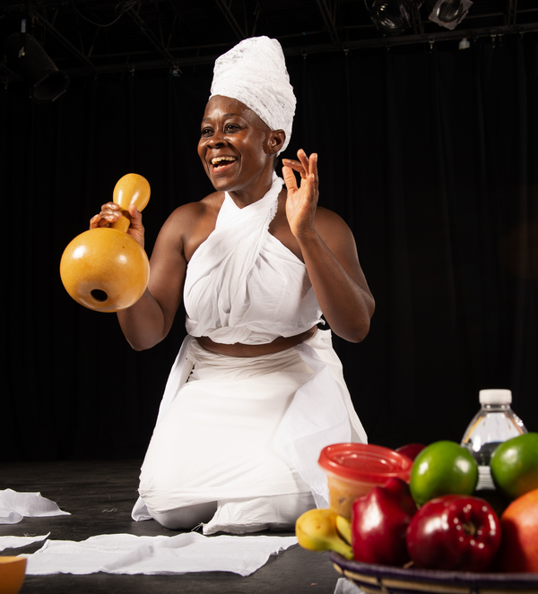 Photograph of Ama Oforiwaa Aduonum mid performance playing a percussive gourd instrument and smiling.