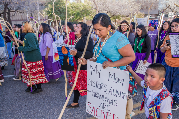Photograph of protesters: women and children wearing colorful clothing and holding signs.