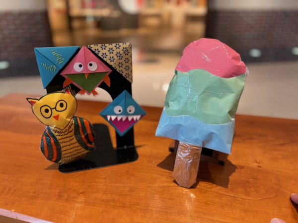 A photo of two paper crafts on a table. One is of a owl and the other is of a popsicle.
