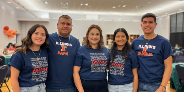 A family of five smiles together at a University of Illinois event while wearing matching navy-blue Illinois and Latinx-themed shirts. The shirts include the words “Illinois Papá,” “Illinois Familia,” and “Latinx.”