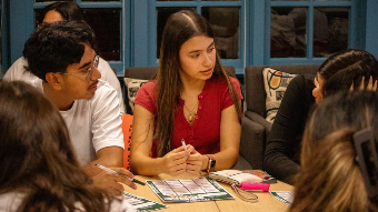 Students gather around a table at La Casa Cultural Latina, engaging in conversation and completing a group activity together. Papers, pens, and activity sheets are spread across the table.