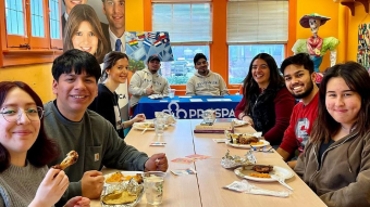 A group of eight college students sit around a long table sharing a meal and smiling toward the camera inside a brightly colored cultural center. Plates of food, drinks, and conversation fill the table, creating a warm, welcoming atmosphere. In the background, additional students sit at an information table with a \\\\\\\\\\\\\\\"Prospera\\\\\\\\\\\\\\\" banner, while colorful artwork, large portraits, international flags, and a Día de los Muertos skeleton decoration add vibrant cultural elements to the space. Large windows bring in natural light, reinforcing the sense of community and belonging.