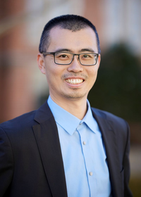 Professor Yuan Yang, wearing glasses, a blue collared shirt, a dark blazer, and smiling