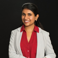Nisha Somnath, wearing a red collared shirt and light grey blazer, smiling