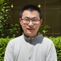 Professor Mengxiao Zhang, wearing a tan zip-up sweater and glasses, in front of a bush