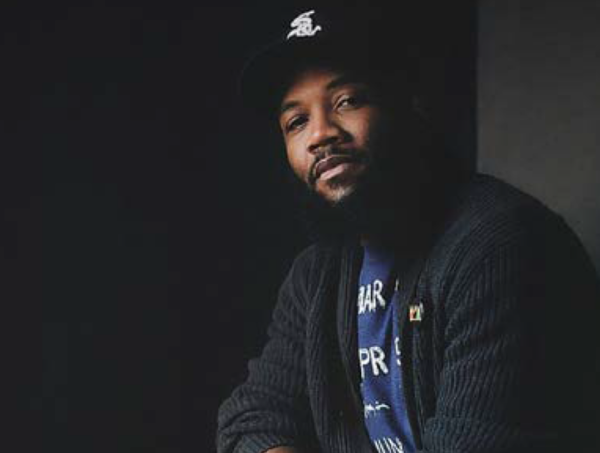 Image of speaker A. D Carson smiling toward the camera. He is wearing a black white sox cap, a blue graphic tee, and a dark grey cardigan over the tee.