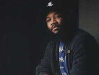 Image of speaker A. D Carson smiling toward the camera. He is wearing a black white sox cap, a blue graphic tee, and a dark grey cardigan over the tee.