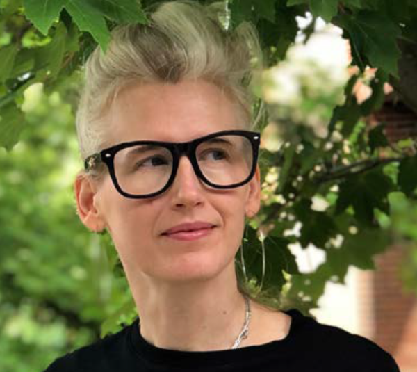 Image of Emily Bevins who is looking to left while smiling. Bevins wears a pair of black glasses, silver hoops, and a black crew neck tee. Bevins also stands in front of a maple tree with green luscious leaves.