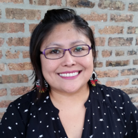 Photo of speaker Yepa-Pappan smiling and looking toward the camera. The artist wears purple prescription glasses and a black and white cat head and polka dot tunic blouse with a low v-cut.