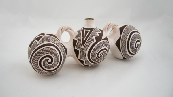 Image of black on white "Triple Canteen" by Juana Leno, has Tulerosa Design. There are three earthenware canteens connected to one another, only the center has an opening for water, in between the canteens there can be seen twisted handles near the top. The canteens are decorated by black staining/paint with one big swirl each accompanied by stripes and zig-zag patterns.