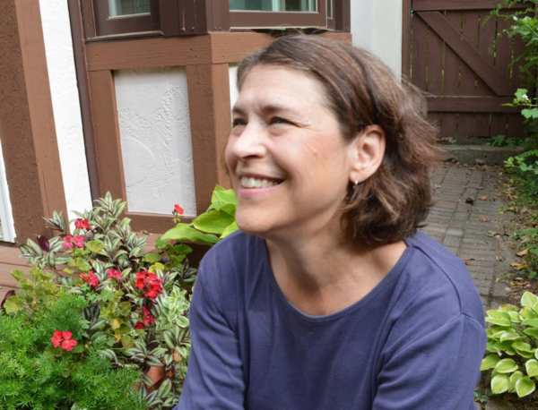 Image of speaker Dede Fairchild Ruggles from an angle. She is smiling a tooth smile. There can be seen red flowers and green leafy plants in the background.