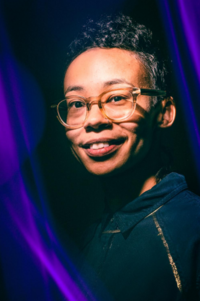 Passionate adult woman with a toothy smile and wearing translucent prescription glasses. There are streaks of violet to her left and upper right.