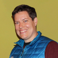 Adult woman in a burgundy shirt and blue puffer jacket vest smiling into the camera with a smile.
