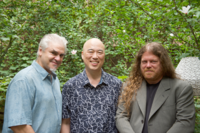 Three adult men looking ahead and smiling in front of green leafed trees.
