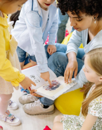 adult holding a book in her lap. She is surrounded by children who are engaging with the words and pictures. Two of the children point at a picture in the book. The kids are young - around 4-5 years old.