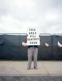 person holding a white sign in front of their face and torso that reads "this area will gentrify soon". Background is a wired fence with a black tarp strewn about the fence.