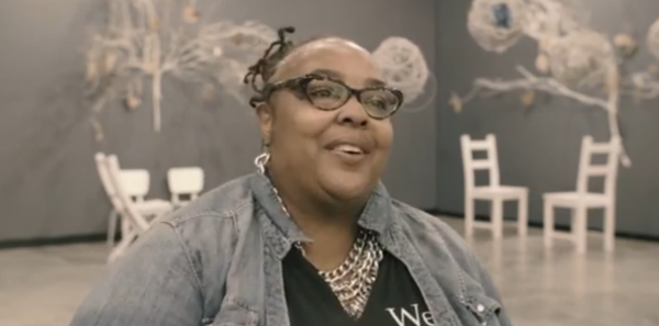 adult woman in a jean jacket and glasses, smiling in front of white wooden chairs and tree-like decor on walls behind her.