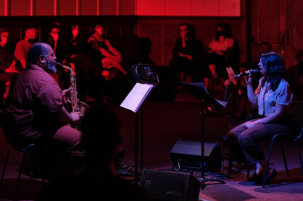 Seated man playing a saxophone to the left facing to the right while a seated woman sings into a microphone facing the saxophonist