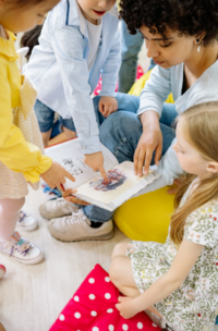 Seated grown woman displaying an open book to toddlers surrounding the woman as they point at images in the book.