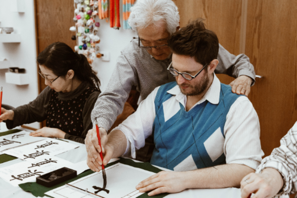 Image of a grown man guiding students through writing kanji with a red brush pen, black ink and posture.