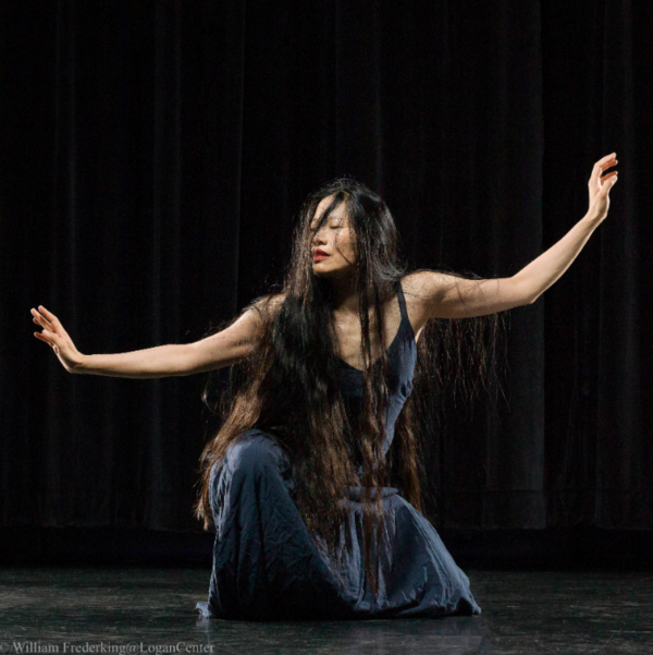 Photo of a woman in a black dress being rained on, squatting mid-dance with her arms extended to her sides.