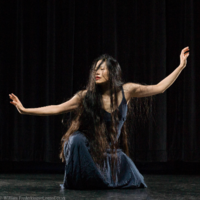 Photo of a woman in a black dress being rained on, squatting mid-dance with her arms extended to her sides.