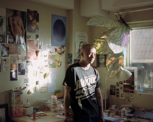 Photo of a tattooed man standing indifferently in a room full of photographs, paper origami, and other crafts materials.