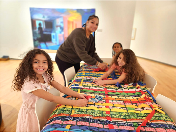 young girls smiling and woman, weaving on a table, in an art gallery