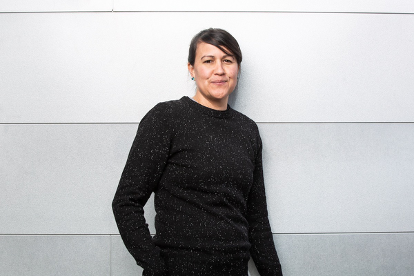 Photo of Natalie Diaz standing in front of a pale gray wall. Diaz is wearing a charcoal grey sweater speckled in white and aqua gemstone stud earrings, and her dark hair is pulled back.