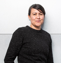 Photo of Natalie Diaz standing in front of a pale gray wall. Diaz is wearing a charcoal grey sweater speckled in white and aqua gemstone stud earrings, and her dark hair is pulled back.