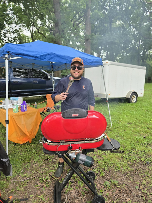 An individual standing at a grill with tongs under a canopy outdoors.