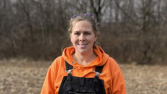 Outdoor headshot of a woman wearing black overalls over an orange hooded sweatshirt.