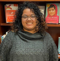 Picture of Joy, with curly hair and glasses, wearing a gray sweater, standing in front of a bookcase showcasing books about leadership.