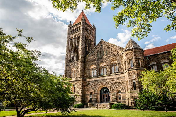 Altgeld Hall stands before a bright blue sky. Clouds are parting so the sun can shine on the sandstone brick of the 2nd oldest building on UIUC's campus. Bright green grass and trees frame the building.