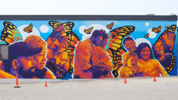 A large, vibrant mural covers the side of a building, featuring several portraits of people and families rendered in bright shades of orange, purple, blue, and yellow. Monarch butterflies with outstretched wings are woven throughout the composition, creating a backdrop behind the figures. The mural extends across the entire wall against a bright blue background with circular white accents.