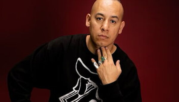 Portrait of artists, Olmeca, wearing a black sweatshirt and turquoise rings, resting one hand beneath his chin against a dark red background.