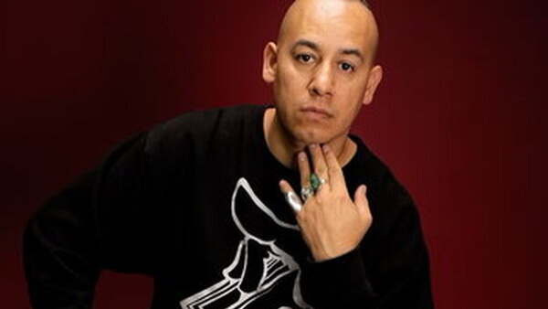Portrait of artist Olmeca, wearing a black sweatshirt and turquoise rings, resting one hand beneath his chin against a dark red background.